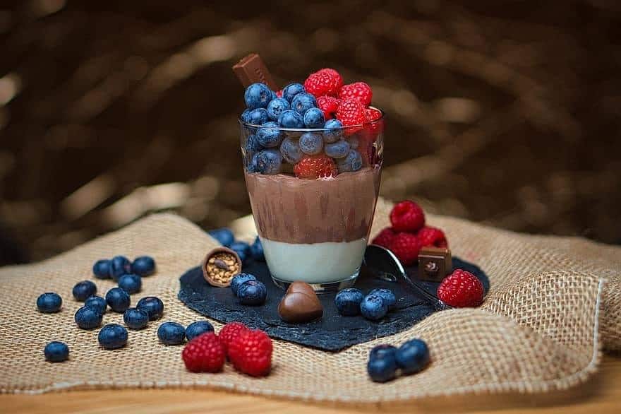 A glass dessert cup sits on a square of burlap and is filled with vanilla and chocolate pudding and topped with blueberries and raspberries.