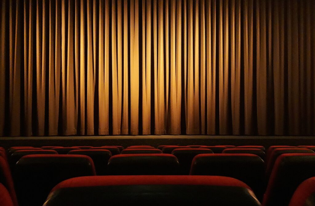 A theatre stage with a gold curtain pulled across it is shown form the audience seating with rich red upholstery.